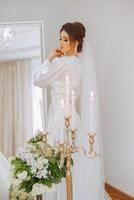 une magnifique brunette la mariée est en train de préparer pour le mariage dans une magnifique boudoir style robe parmi Frais fleurs et élégant chandeliers contre une miroir Contexte. mariage portrait, photo. photo