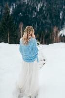 arrière voir, verticale photo. une mignonne mariée, enveloppé dans une bleu plaid, est attendre pour le jeune marié contre le Contexte de une hiver paysage. photo