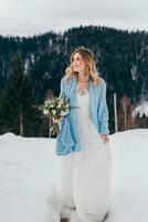 portrait de une mignonne la mariée dans une blanc robe avec une bouquet de fleurs dans sa mains, enveloppé dans une bleu plaid, attendre pour le jeune marié contre le Contexte de une hiver paysage de enneigé montagnes. photo