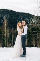 toute la longueur portrait de le la mariée et jeune marié contre le Contexte de une pin forêt. le la mariée dans une blanc mariage robe, le jeune marié dans une blanc chemise et gilet. hiver mariage concept. photo