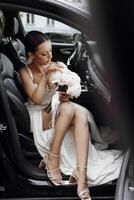 une aux cheveux noirs la mariée est assis dans une moderne noir voiture sur votre mariage journée avec une bouquet de fleurs. portrait de le la mariée. blanc robe, longue voile photo