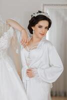 Matin de le la mariée avant le mariage. une magnifique Jeune femme avec une voile et une magnifique coiffure dans une blanc peignoir près sa mariage robe sur une mannequin. professionnel se maquiller. dernier les préparatifs. photo