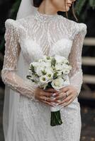 le la mariée dans une blanc mariage robe brodé avec des pierres et perles détient une mariage bouquet de des roses et verdure. élégant mariage bouquet. photo