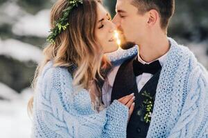 portrait de le la mariée et jeune marié contre le Contexte de une hiver forêt, magnifique lumière derrière. le la mariée et jeune marié sont enveloppé dans une bleu couverture. une seconde avant le baiser. hiver mariage. photo