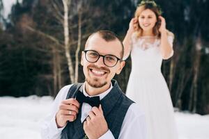 portrait de le jeune marié dans le premier plan dans une blanc chemise et gilet et une arc attacher contre le Contexte de une pin forêt. hiver mariage concept. photo