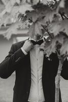 le jeune marié dans une noir costume ajuste le sien veste, pose contre le Contexte de une vert arbre. mariage noir et blanc portrait. photo