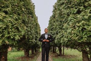 le jeune marié dans une noir costume ajuste le sien veste, pose contre le Contexte de une vert arbre. mariage portrait. photo