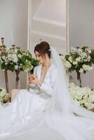 une magnifique brunette la mariée est avoir prêt pour le mariage dans une magnifique boudoir style tenue séance sur une chaise parmi Frais fleurs et élégant chandeliers. mariage portrait, photo. photo