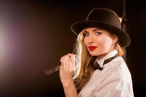 Jeune femme séduisante blonde en chemise blanche, papillon, chapeau noir avec microphone chantant sur scène photo