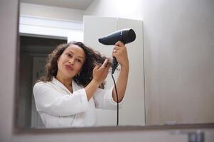 multi ethnique brunette, attrayant Jeune femme dans blanc peignoir de bain, séchage cheveux avec cheveux séchoir dans salle de bains à Hôtel Accueil photo