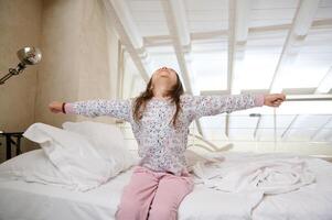 content peu enfant fille élongation bras vers le haut tandis que séance sur lit, en dessous de blanc couverture tandis que veille en haut dans le Matin photo