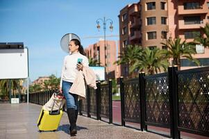 plein longueur portrait de une femme avec Jaune bagage, à la recherche de côté, en portant mobile téléphone tandis que en marchant dans ville rue photo