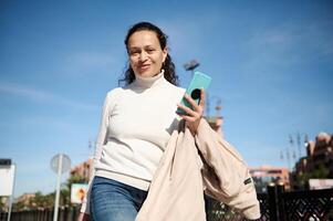 magnifique femme en portant mobile téléphone tandis que ambulant le ville rue, souriant joliment à la recherche à le caméra photo
