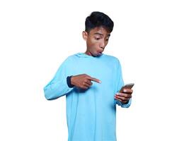 Jeune asiatique homme surpris à la recherche à intelligent téléphone portant bleu T-shirt, isolé blanc Contexte. photo