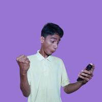 asiatique homme content plus de difficile travail la victoire, la gauche main en portant intelligent téléphone isolé violet Contexte. photo