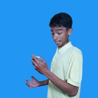 Jeune asiatique homme surpris à la recherche à intelligent téléphone isolé bleu Contexte. portant une Jaune T-shirt photo