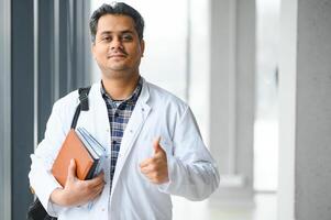 portrait de une Jeune Indien Masculin médical étudiant dans une blanc manteau agitant photo