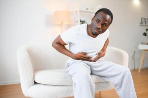 africain américain gars ayant estomac mal après en mangeant émouvant douloureux estomac Souffrance de douleur séance sur canapé à maison. photo