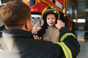 une pompier spectacles le sien travail à le sien Jeune fils. une garçon dans une sapeurs pompiers casque photo