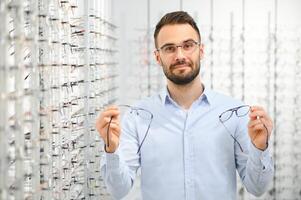 choisir des lunettes concept. portrait de Beau barbu gars cueillette Nouveau spécifications à optique boutique, à la recherche à miroir photo