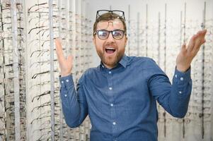 satisfait client. vue de content Jeune Masculin client portant Nouveau lunettes, permanent près grille et vitrine avec lunettes. souriant homme en essayant sur lunettes photo