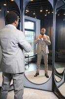 vente, achats, mode, style et gens concept - africain homme choisir et en essayant veste ou costume sur et à la recherche à miroir dans centre commercial ou Vêtements boutique photo