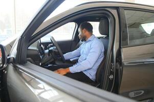 voiture propriétaire. joyeux afro gars souriant, séance dans Nouveau voiture conduite de concession magasin photo