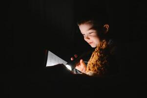 une garçon de 5-6 ans vieux est en train de lire une livre dans le soir dans le foncé en dessous de une couverture avec une jouet ours. photo