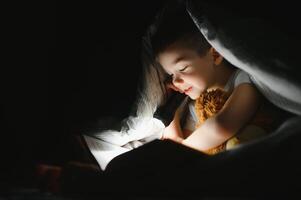une garçon de 5-6 ans vieux est en train de lire une livre dans le soir dans le foncé en dessous de une couverture avec une jouet ours. photo