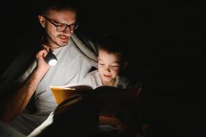 content famille en train de lire heure du coucher récit en dessous de couverture dans soir. père et fils dépenser temps ensemble. du père journée photo