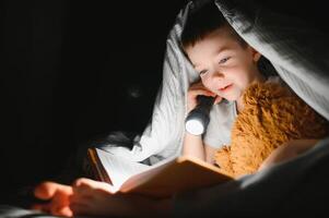 enfant en train de lire livre dans lit. des gamins lis à nuit. peu garçon avec Fée conte livres dans chambre . éducation pour Jeune les enfants. heure du coucher récit dans le soir. mignonne enfant en dessous de couverture dans foncé pièce avec lampe photo