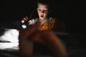 portrait de mignonne peu garçon en train de lire dans lit avec lampe de poche dans foncé chambre, profiter contes de fées. photo