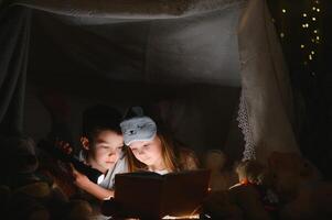 peu des gamins impliquant dans en train de lire incroyable livre. elles ou ils mensonge dans agréable jouet tente dans salle de jeux. garçon en portant lampe de poche dans main. photo