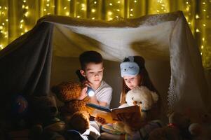 deux les enfants avec lampe de poche lis une livre en dessous de une couverture comme une tente. photo