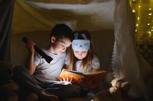 peu des gamins impliquant dans en train de lire incroyable livre. elles ou ils mensonge dans agréable jouet tente dans salle de jeux. garçon en portant lampe de poche dans main. photo