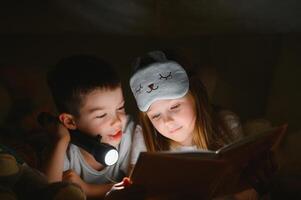 en train de lire et famille Jeux dans enfants tente. garçon et fille avec livre et lampe de poche avant Aller à lit. photo