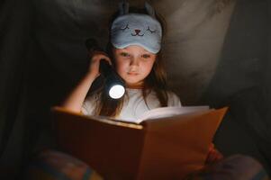 enfant fille en train de lire avec livre et lampe de poche et nounours ours dans tente. avant Aller à lit. photo
