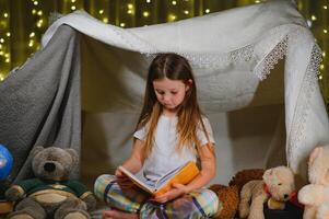 peu fille sont en train de lire une livre avec lampes de poche dans tente. content fille en jouant à maison. marrant charmant enfant ayant amusement dans pièce photo