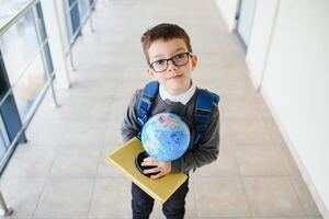 content souriant garçon dans des lunettes est Aller à école pour le premier temps. enfant avec école sac et livre dans le sien main. enfant à l'intérieur de le classe pièce . retour à école photo