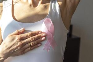 coup de le femme contre le blanc mur dans le blanc Haut avec rose ruban, comme une symbole de une Sein cancer conscience, performant soi examen de le seins, à la recherche pour anomalies. concept photo