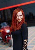 une élégant roux posant suivant à une lisse moto. une femme avec rouge cheveux permanent suivant à une moto photo