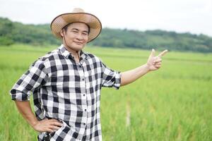 Beau asiatique homme agriculteur est à paddy champ, porte chapeau et plaid chemise, mettre chapeau sur taille, point doigt en haut. concept, agriculture profession. thaïlandais agriculteur. travail avec la nature. biologique agriculture. photo