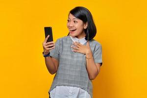 joyeuse jeune femme asiatique regardant un téléphone portable et gardant les paumes sur la poitrine sur fond jaune photo