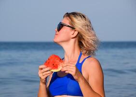 une blond dans des lunettes de soleil mange une pastèque par le mer. une juteux pastèque dans le mains de une femme photo