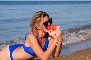 le blond dans des lunettes de soleil mensonges sur le plage près le vagues et mange une pastèque. une juteux pastèque dans le mains de une femme. photo