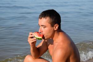 une homme est en mangeant une pastèque séance sur le plage. une mûr pastèque dans le mains de une homme photo