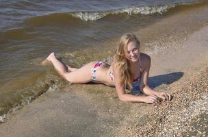 blond fille dans une bikini mensonge sur le plage et le vagues éclaboussure sur il. magnifique Jeune femme dans une coloré bikini sur mer Contexte photo