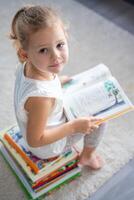 portrait de peu fille est séance sur empiler de enfants livres et feuilletage par une livre avec Fée contes photo