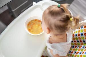 mignonne bébé fille bambin séance dans le haute chaise et en mangeant sa le déjeuner soupe à Accueil cuisine. photo