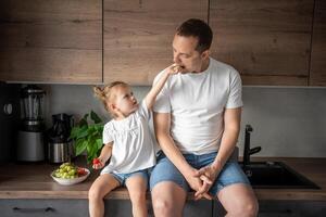 mignonne peu fille et sa Beau papa sont en mangeant fruit dans moderne cuisine. en bonne santé alimentaire. photo